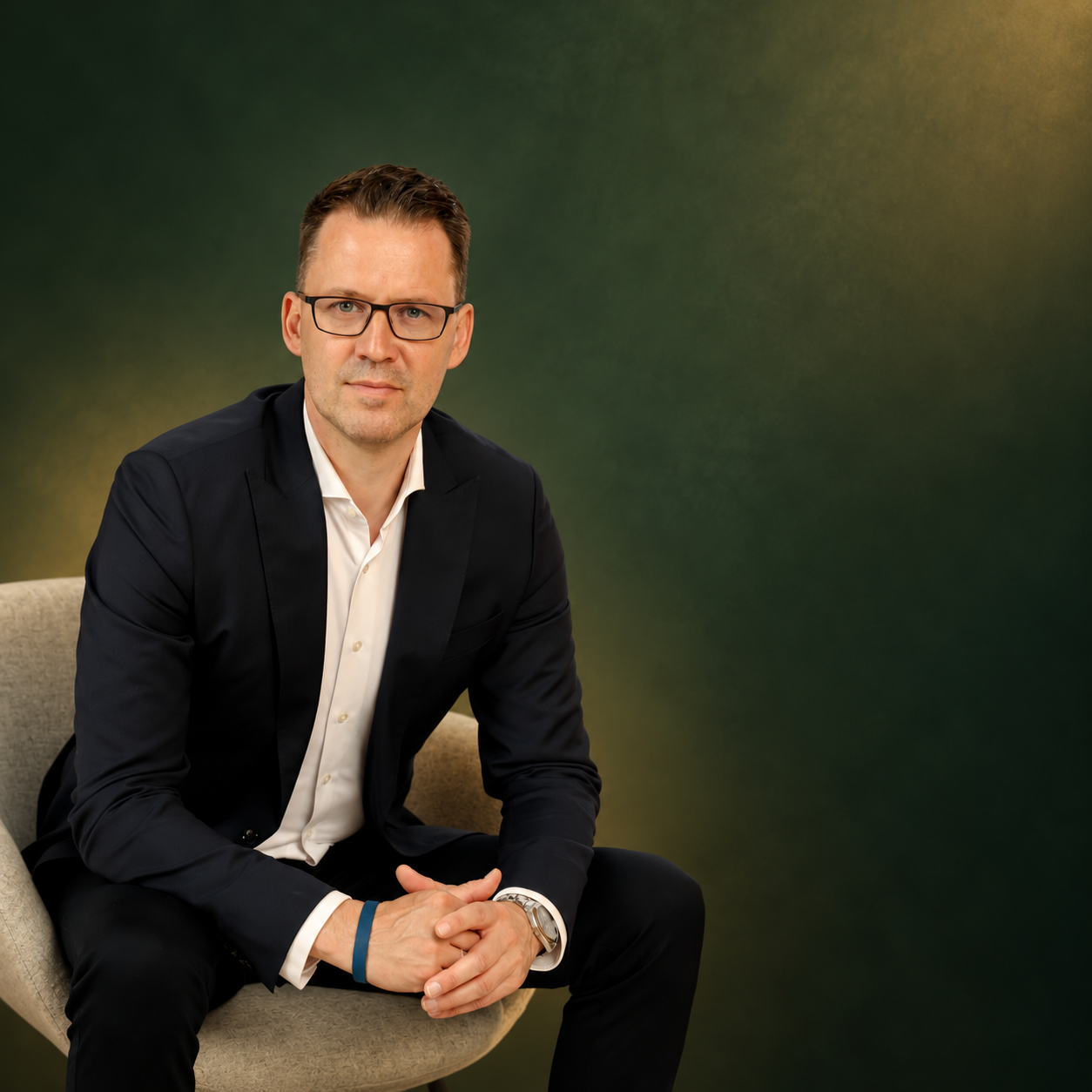 Danny Kukulies sitzt im dunklen Anzug auf einem Sessel vor dunkelgrünem Hintergrund.